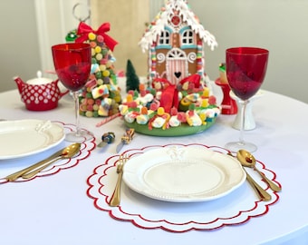 May include: A festive table setting with red wine glasses, white plates, and gold cutlery. A gingerbread house and candy decorations create a holiday theme. Red scalloped placemats and a white tablecloth complete the Christmas scene.
