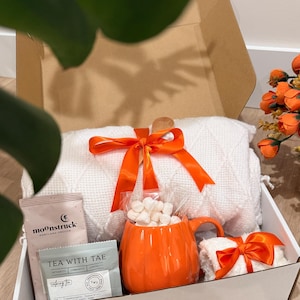 May include: A gift box containing a white textured throw blanket tied with an orange ribbon, an orange pumpkin-shaped ceramic mug filled with white marshmallows, and two tea packets labeled Moonstruck and Tea With Tae with peppermint ingredients.