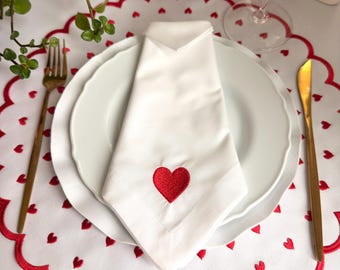 May include: A white cloth napkin featuring a single embroidered red heart, folded and placed on a white dinner plate. The plate rests on a round white placemat with a scalloped red edge and a pattern of small red hearts, with gold cutlery.