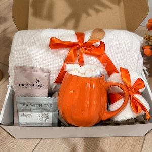 May include: A gift box containing a white textured throw blanket tied with an orange ribbon, a bright orange pumpkin-shaped ceramic mug filled with white marshmallows, a Moonstruck chocolate bar, and a Tea With Tae tea bag in a white box.