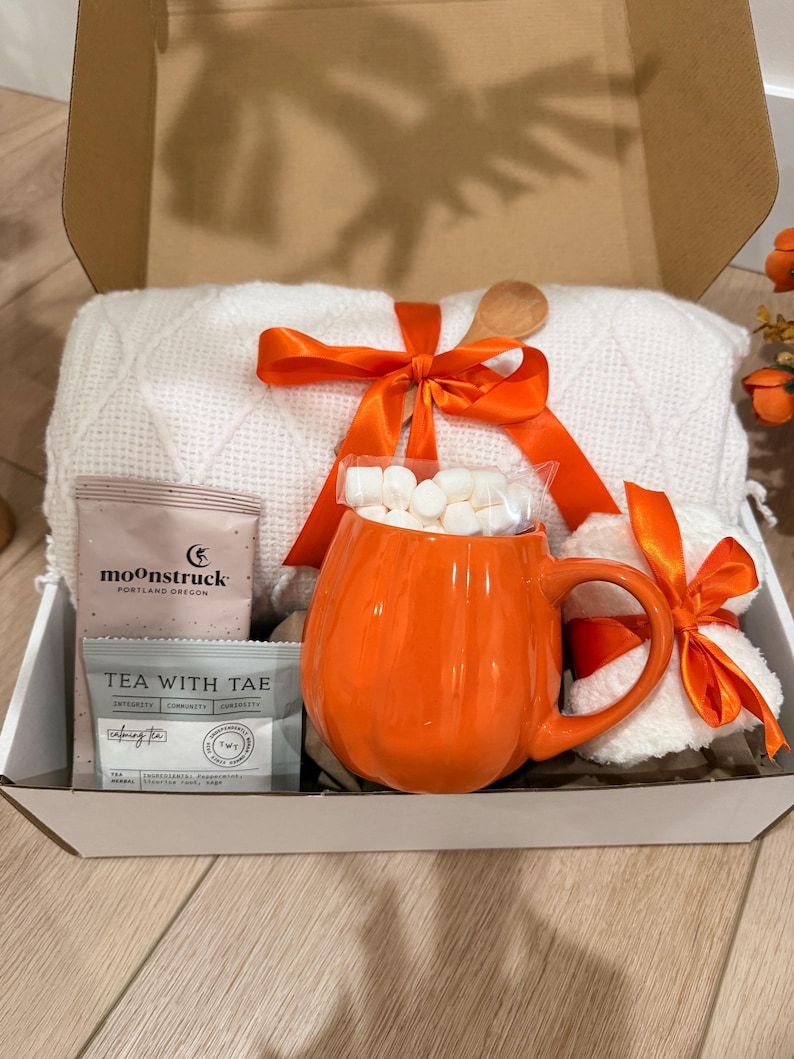 May include: A gift box containing a white textured throw blanket tied with an orange ribbon, a bright orange pumpkin-shaped ceramic mug filled with white marshmallows, a Moonstruck chocolate bar, and a Tea With Tae tea bag in a white box.