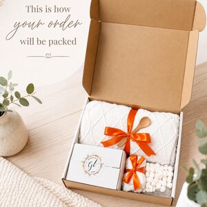 May include: An open cardboard shipping box containing a white textured knit blanket tied with an orange ribbon, a small wooden spoon, a white gift box with a circular logo, a small white towel with an orange ribbon, and white marshmallows.