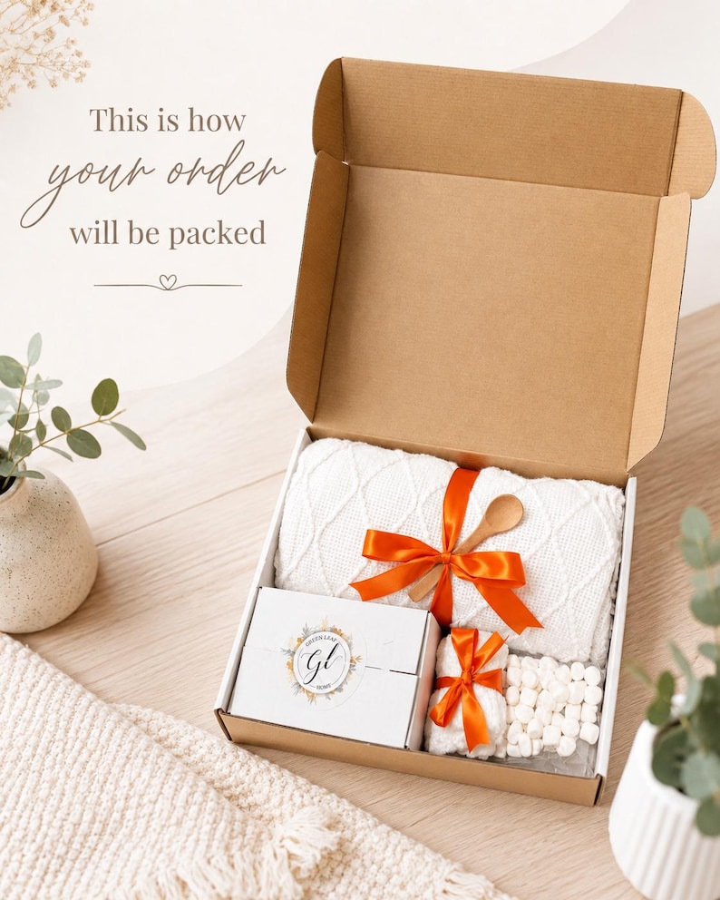 May include: An open cardboard shipping box containing a white textured knit blanket tied with an orange ribbon, a small wooden spoon, a white gift box with a circular logo, a small white towel with an orange ribbon, and white marshmallows.