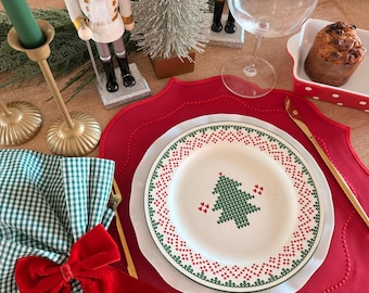 May include: A white ceramic plate with a green knitted-style Christmas tree pattern and red heart accents, centered on a red scalloped placemat. The setting includes a green and white gingham napkin with a red velvet bow and gold cutlery.