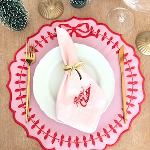 May include: A light pink scalloped placemat with red embroidered holly and bow details, a white dinner plate, a light pink napkin with red Merry Christmas embroidery, a gold bow napkin ring, a gold fork, and a gold knife on a wooden surface.