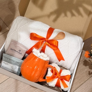 May include: A gift box containing a white cable-knit blanket tied with an orange ribbon, a pumpkin-shaped orange mug filled with white marshmallows, a small white plush item with an orange ribbon, and two tea packets labeled Moonstruck and Tea With Tae.