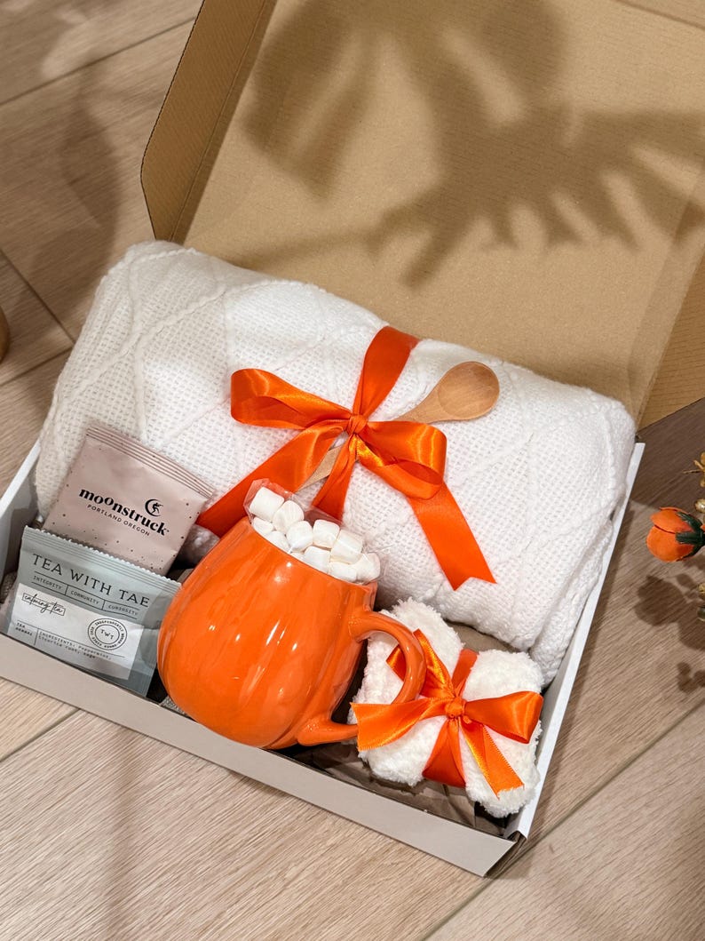 May include: A gift box containing a white cable-knit blanket tied with an orange ribbon, a pumpkin-shaped orange mug filled with white marshmallows, a small white plush item with an orange ribbon, and two tea packets labeled Moonstruck and Tea With Tae.