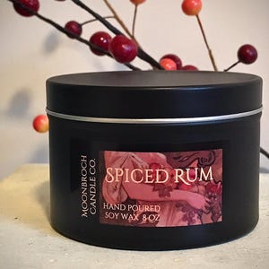 May include: A black candle tin with a silver rim and a label that reads "Spiced Rum." The label features an illustration and the text "Hand Poured Soy Wax 8 oz." Red berries and branches are in the background.