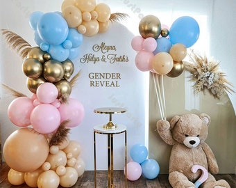 May include: A gender reveal party setup with a white backdrop decorated with gold and pink balloons, a teddy bear, and a sign that reads "Aleyna, Hulya & Fatih's Gender Reveal".