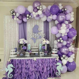 Peut inclure: Une arche de ballons violette et argentée avec une silhouette de sirène portant l'inscription "Brianna" au-dessus d'une table avec une nappe à volants violette et deux décorations en forme d'hippocampe.