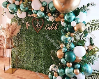 May include: A decorative balloon arch in shades of gold, teal, and white, with greenery and a backdrop of faux green foliage. The arch includes the names "Re & Caleb" in wooden letters, perfect for a wedding or party.