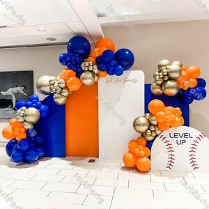 May include: A baseball-themed party backdrop with blue, orange, and gold balloons. The backdrop features a white panel with the text "We Are Family" and a white baseball with the text "Level Up".