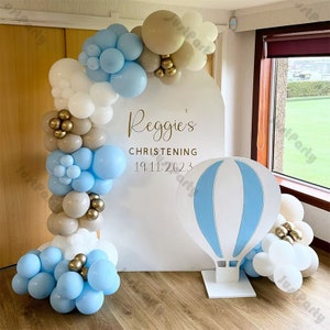 Pode incluir: Um fundo branco com o texto "Reggie's Christening 19.11.2023" em letras douradas. Um recorte de balão de ar quente azul e branco está na frente do fundo. Um arco de balões feito de balões brancos, azuis e dourados está no lado esquerdo do fundo.