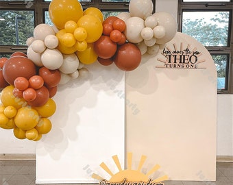 May include: A white and beige backdrop with a balloon arch made of yellow, brown, and cream balloons. The arch is decorated with a wooden sign that says "Here comes the sun THEO TURNS ONE". A yellow sun cutout with the text "@greydesigns.decor" is in the foreground.