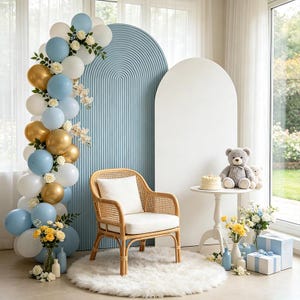 May include: A rattan armchair with a white cushion sits before a blue arched panel and a white arched panel. A balloon arch in blue, white, and gold features floral accents. A small white table holds a cake and a gray teddy bear.