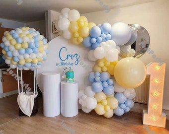 May include: A white backdrop with the name "Cruz" and "1st Birthday" written on it. A balloon arch made of blue, white, and yellow balloons. A white cake with blue and green decorations. Two white pillars. A white hot air balloon made of blue and yellow balloons. A light-up number one.