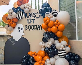 May include: A Halloween-themed balloon arch with orange, black, white, and striped balloons. The arch surrounds a sign that reads "Kai's 1st Booday" with a ghost cutout. The balloons are arranged in clusters and arches.