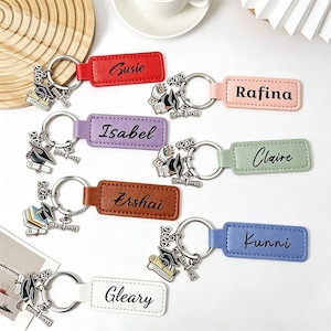 May include: A collection of personalized graduation keychains. Each keychain features a leather tag with a name, such as "Susie" or "Rafina", and a metal charm with a graduation cap and the year "2026". The tags come in various colors.