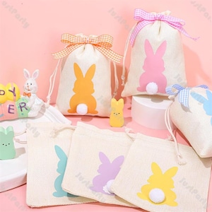 Puede incluir: Bolsas con cordón temáticas de Pascua con siluetas de conejos en naranja, rosa, azul y amarillo. Cada bolsa tiene una cola de pompón blanco y está atada con una cinta. También hay figuras decorativas de Pascua.
