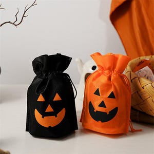 Op de afbeelding: Twee Halloween-snoepzakjes met trekkoord. Eén is zwart met een oranje pompoengezicht en de andere is oranje met een zwart pompoengezicht. De zakken zijn gemaakt van een non-woven stof.