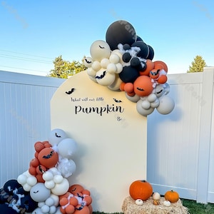 Könnte beinhalten: Eine Halloween-Ballon-Dekoration mit orangefarbenen, schwarzen und weißen Luftballons und einem Schild mit der Aufschrift "What will our little Pumpkin Be...". Kürbisse und Heuballen befinden sich im Vordergrund, mit einem weißen Zaun und blauem Himmel im Hintergrund.