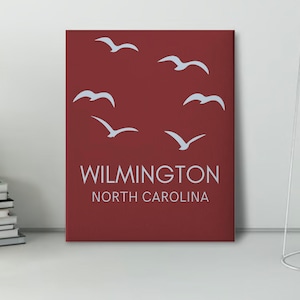 Könnte beinhalten: Ein roter Leinwanddruck mit einer weißen Silhouette von Möwen, die im Kreis fliegen. Der Text "WILMINGTON NORTH CAROLINA" ist in weiß am unteren Rand des Drucks gedruckt.