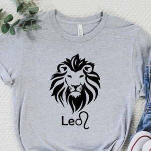 Może przedstawiać: Szary T-shirt z czarnym nadrukiem lwa i tekstem "Leo" wraz z astrologicznym symbolem Lwa.