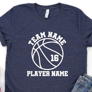 May include: Navy blue t-shirt with a white basketball graphic. The graphic includes the text "TEAM NAME", a basketball, the number "16", and the text "PLAYER NAME". The shirt is paired with denim shorts and white sneakers.