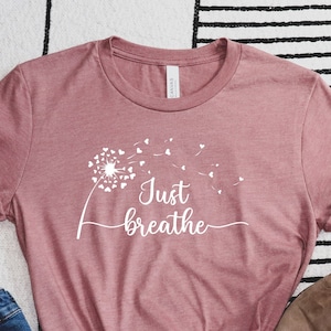 K&ouml;nnte beinhalten: Ein mauvefarbenes T-Shirt mit einem wei&szlig;en L&ouml;wenzahnmuster und dem Text "Just Breathe" in einer kursivschrift.