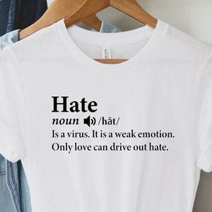May include: White t-shirt with black text that reads "Hate noun /hāt/ Is a virus. It is a weak emotion. Only love can drive out hate."