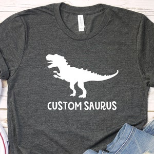 May include: A dark gray t-shirt with a white dinosaur silhouette and the text "CUSTOM SAURUS" printed on the front. The shirt is laid out on a white wooden surface with a pair of white sneakers and blue jeans.
