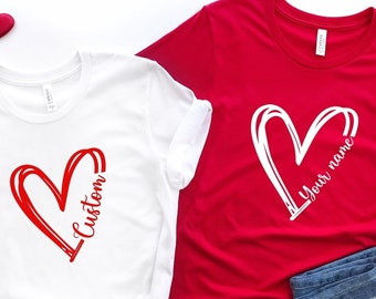 May include: Two t-shirts with a red heart design. One shirt is white with the word "Custom" written inside the heart. The other shirt is red with a white heart and the text "Your name" written inside the heart.