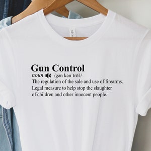 May include: A white t-shirt with the words "Gun Control" printed in black. The definition of gun control is also printed on the shirt. The shirt is hanging on a wooden hanger.