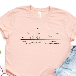 May include: A peach-coloured t-shirt with a black graphic of a sunset over the ocean with birds flying above. The t-shirt is paired with light blue jeans and white trainers.