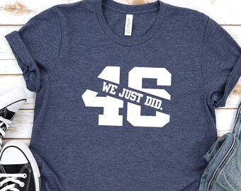 Op de afbeelding: Een marineblauw t-shirt met witte tekst die "WE JUST DID." zegt en het nummer "46" in een groot wit lettertype. Het shirt ligt op een wit houten oppervlak met een paar zwarte sneakers en een blauwe jeans.
