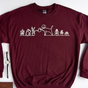 May include: A maroon sweatshirt with a white outline of a dog watering a row of potted plants. The dog is holding a watering can and the plants are various cacti and succulents.
