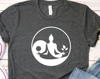 Op de afbeelding: Een donkergrijs T-shirt met een wit yin yang symbool met een persoon die erin mediteert. Het symbool heeft aan de ene kant een blad en aan de andere kant een cirkel.