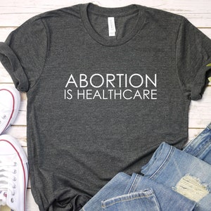 May include: A dark gray t-shirt with the text "ABORTION IS HEALTHCARE" printed in white on the front. The shirt is laid flat on a white wooden surface with a pair of white sneakers, blue jeans, and a potted plant.