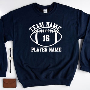 May include: Navy blue sweatshirt with a white football graphic and the text "TEAM NAME 16 PLAYER NAME".