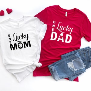 May include: Two white t-shirts with black text and a shamrock design. The text on the white shirts reads "One Lucky Mom" and "One Lucky Dad". A red t-shirt with white text and a shamrock design reads "One Lucky Dad".