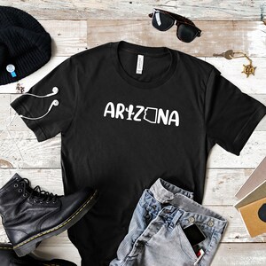 May include: Black t-shirt with white text that says "ARIZONA" with the state outline inside the "A". The shirt is laid flat on a woodgrain surface with a beanie, sunglasses, jeans, a phone, and notebooks.