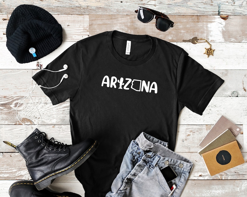 May include: Black t-shirt with white text that says "ARIZONA" with the state outline inside the "A". The shirt is laid flat on a woodgrain surface with a beanie, sunglasses, jeans, a phone, and notebooks.