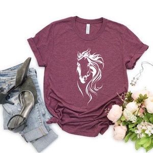 May include: A maroon t-shirt with a white silhouette of a horse's head. The shirt is laid flat on a white background with a pair of jeans and a bouquet of flowers.