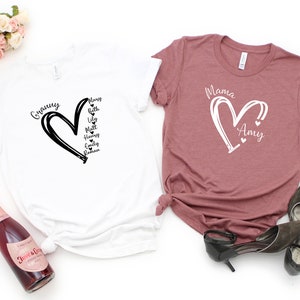 K&ouml;nnte beinhalten: Zwei T-Shirts mit einem Herzdesign und Namen im Herzen. Das wei&szlig;e T-Shirt sagt "Granny" und hat die Namen "Mary, Beth, Lily, Matt, Henry, Emily, Roman" im Herzen. Das rosa T-Shirt sagt "Mama" und hat den Namen "Amy" im Herzen.