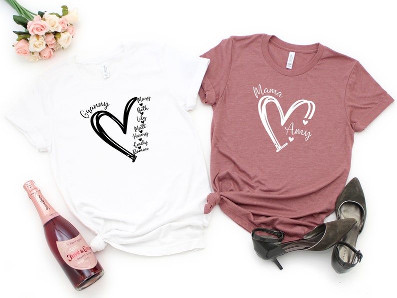 K&ouml;nnte beinhalten: Zwei T-Shirts mit einem Herzdesign und Namen im Herzen. Das wei&szlig;e T-Shirt sagt "Granny" und hat die Namen "Mary, Beth, Lily, Matt, Henry, Emily, Roman" im Herzen. Das rosa T-Shirt sagt "Mama" und hat den Namen "Amy" im Herzen.