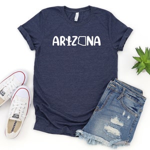 May include: A navy blue t-shirt with the word "Arizona" printed in white with a stylized outline of the state of Arizona. The shirt is shown with a pair of white sneakers, denim shorts, and a small potted plant.