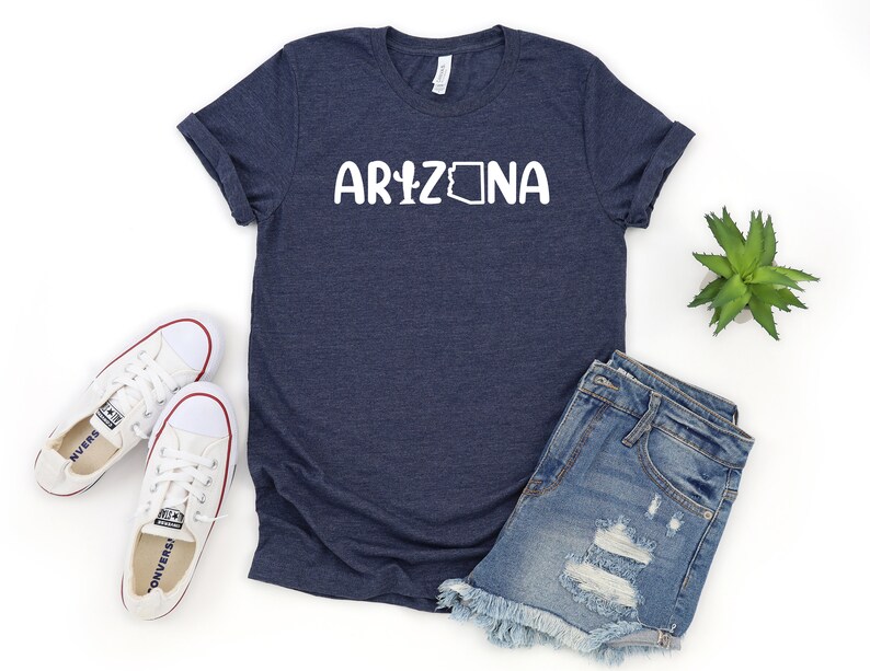May include: A navy blue t-shirt with the word "Arizona" printed in white with a stylized outline of the state of Arizona. The shirt is shown with a pair of white sneakers, denim shorts, and a small potted plant.