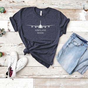 May include: A dark blue t-shirt with a white airplane graphic and the text "AIRPLANE MODE". The shirt is laid flat on a white wooden surface with a pair of white sneakers and a pair of light blue jeans.