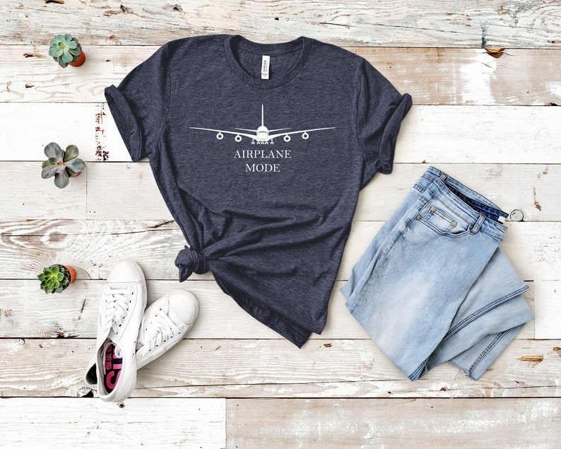 May include: A dark blue t-shirt with a white airplane graphic and the text "AIRPLANE MODE". The shirt is laid flat on a white wooden surface with a pair of white sneakers and a pair of light blue jeans.