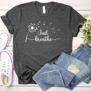 K&ouml;nnte beinhalten: Ein dunkelgraues T-Shirt mit einem wei&szlig;en L&ouml;wenzahnmuster und dem Text "Just breathe". Das Shirt liegt auf einer wei&szlig;en Holzoberfl&auml;che mit einem Paar wei&szlig;en Turnschuhen und einer blauen Jeans.